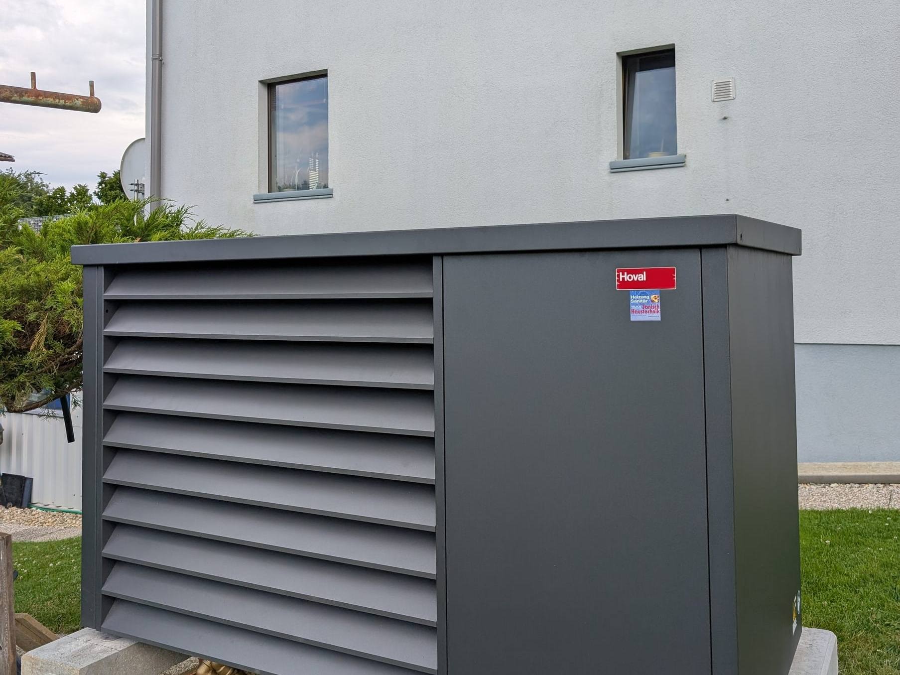 Anthrazitfarbene Hoval-Wärmepumpen-Außeneinheit mit Lamellenverkleidung auf Betonbalken vor einem Wohnhaus
