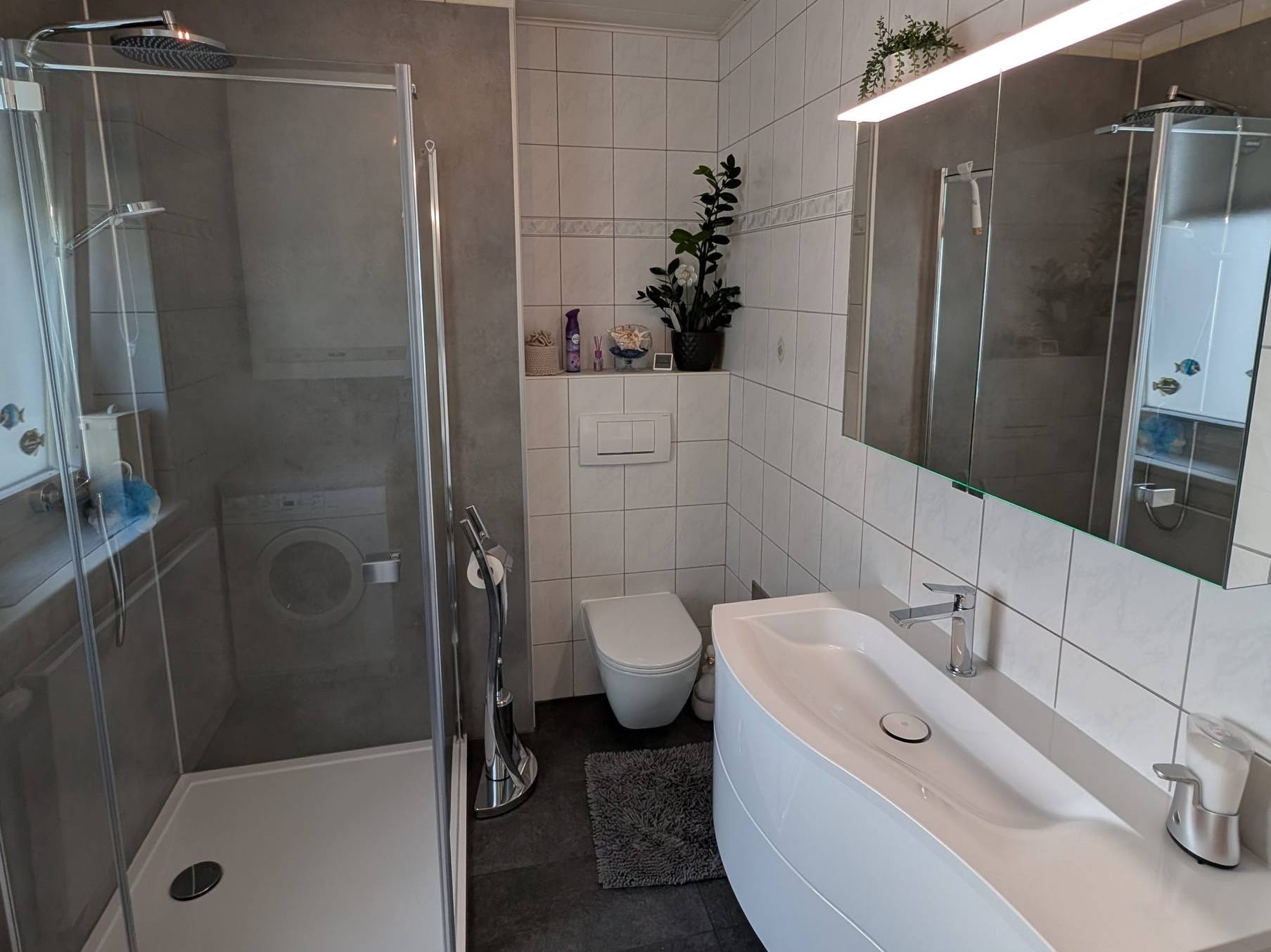 Renoviertes Bad mit Eckdusche, wandhängendem WC, weißem Waschtisch und beleuchtetem Spiegelschrank