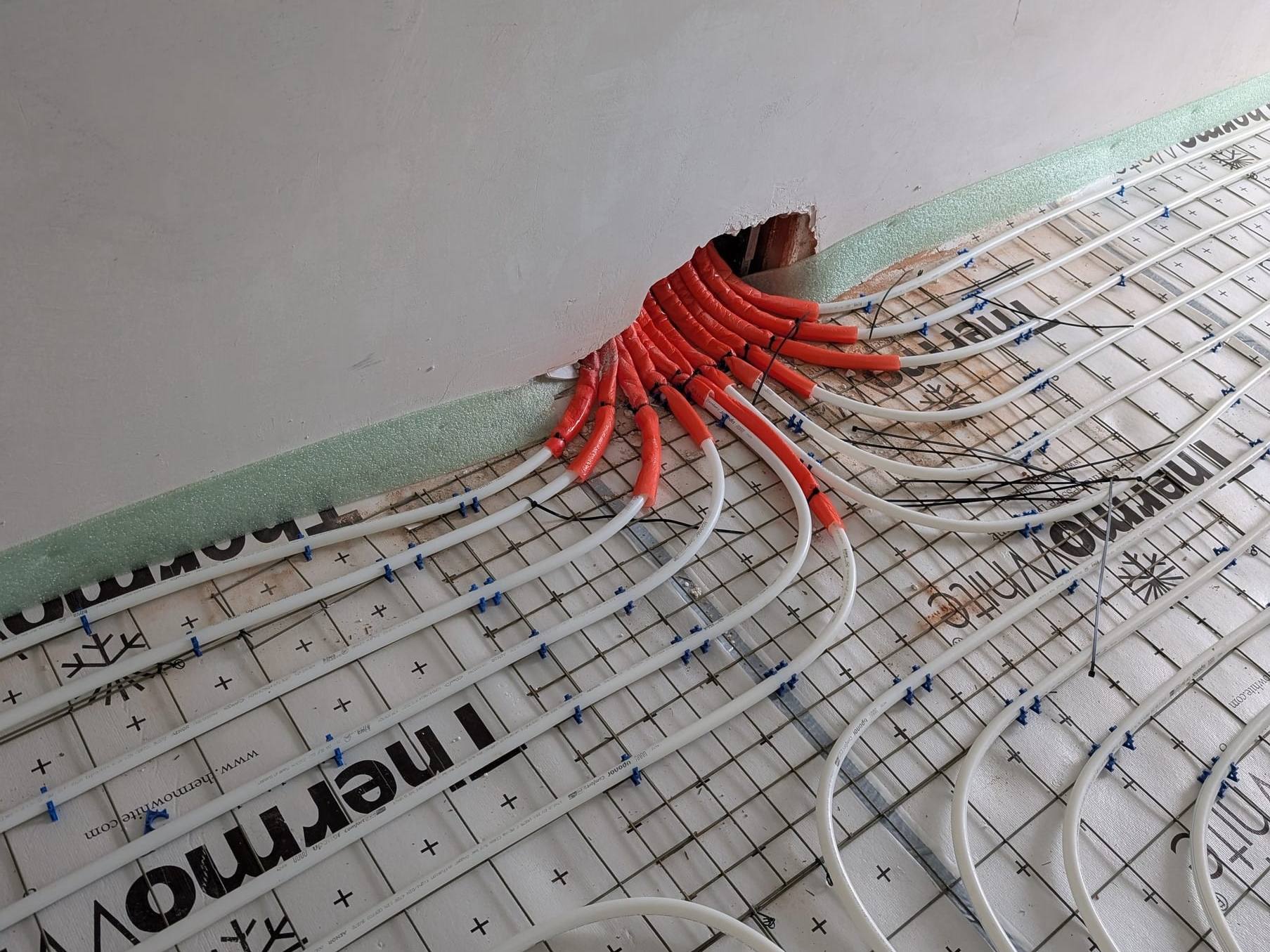 Fußbodenheizungsrohre mit roten Schutzhülsen führen gebündelt durch eine Wandöffnung