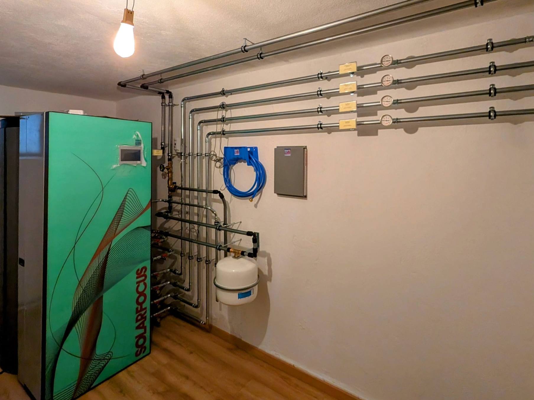 Heizraum mit grünem Solarfocus-Gerät, beschrifteten Heizkreisleitungen und weißem Ausdehnungsgefäß