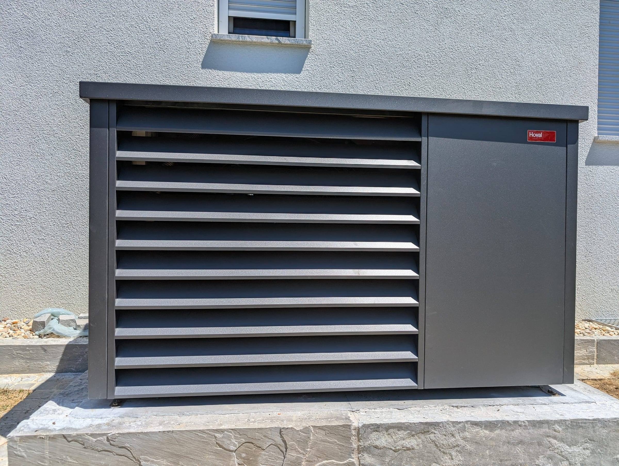 Anthrazitfarbene Hoval-Wärmepumpe mit Lamellenfront auf Natursteinsockel vor Hauswand