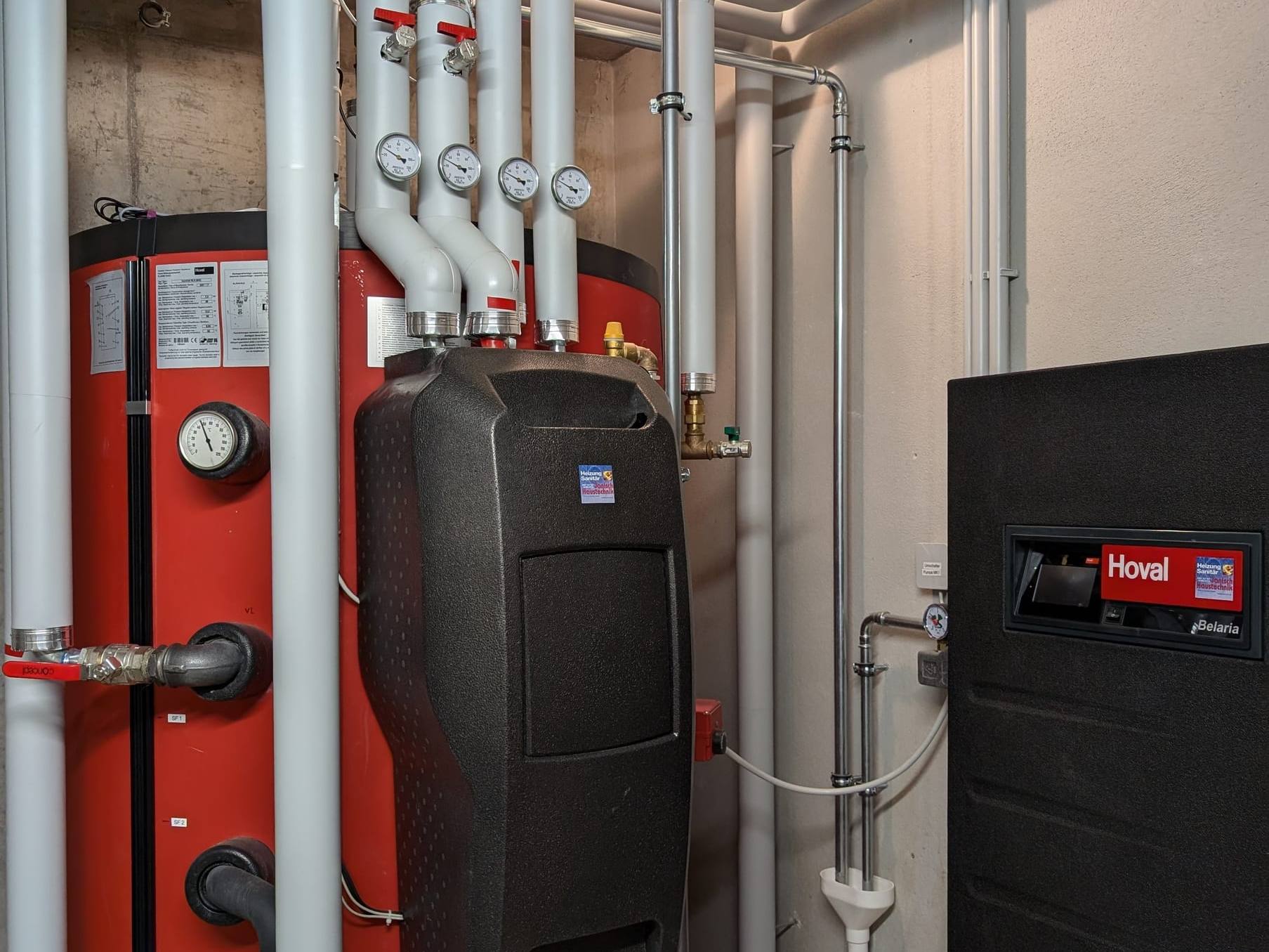 Hoval-Belaria-Wärmepumpe neben rotem Speicher mit weiß gedämmten Rohrleitungen im Technikraum