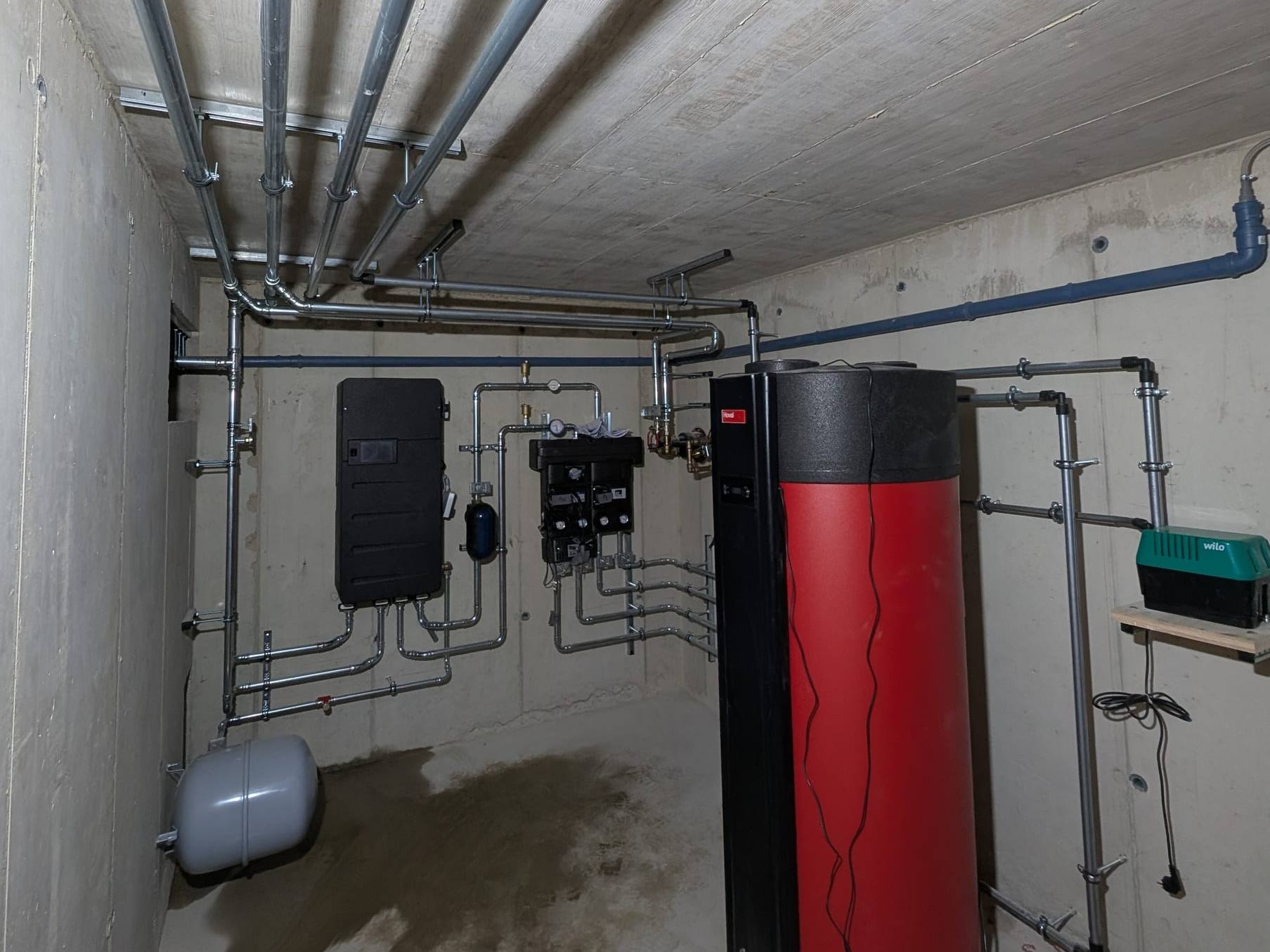 Technikraum mit rot-schwarzem Hoval-Speicher, Pumpengruppen und verzinkten Leitungen im Betonkeller