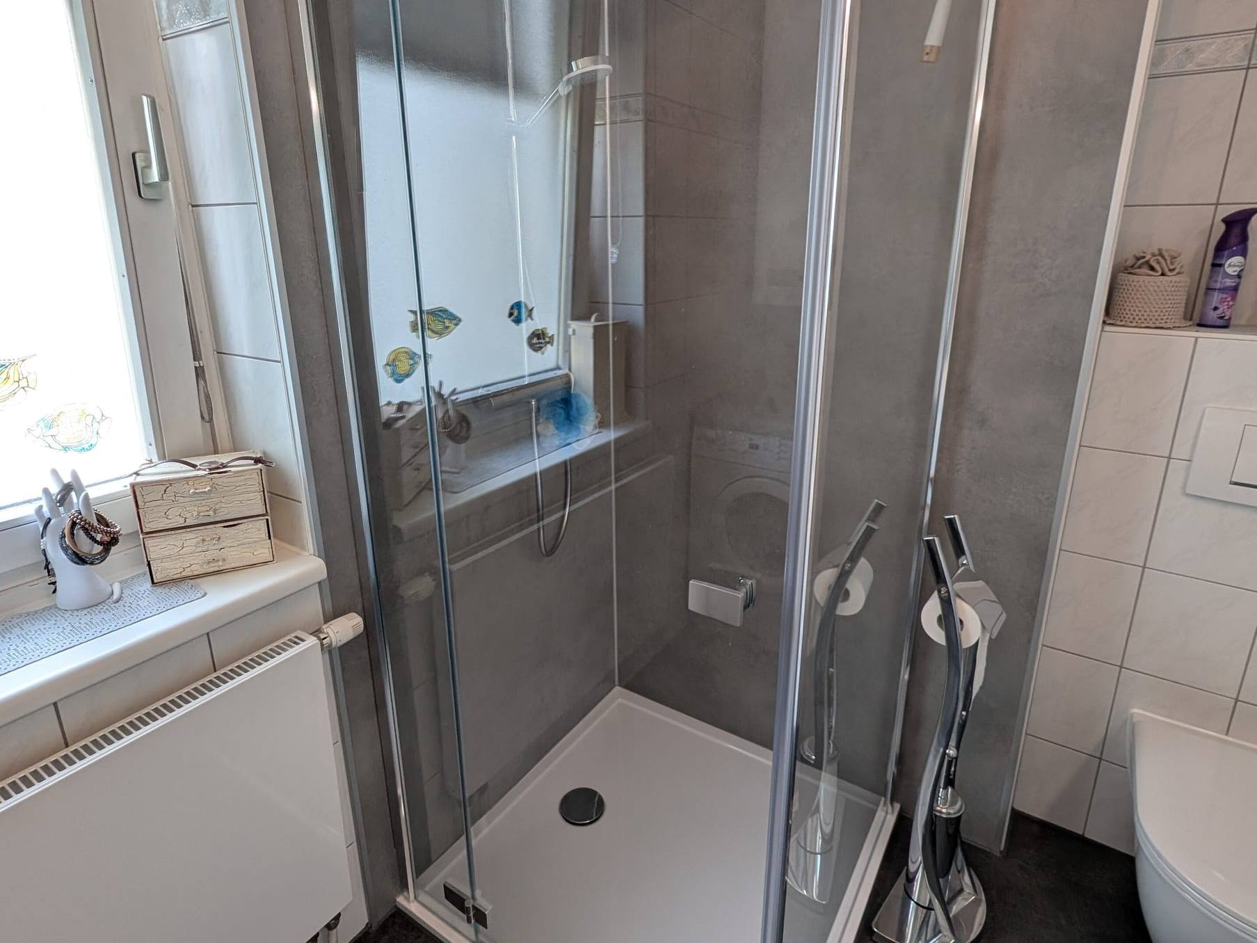 Eckdusche mit Glastür, Regenbrause, weißer Duschtasse und grauer Rückwand aus Respatex-Platten neben einem Fenster