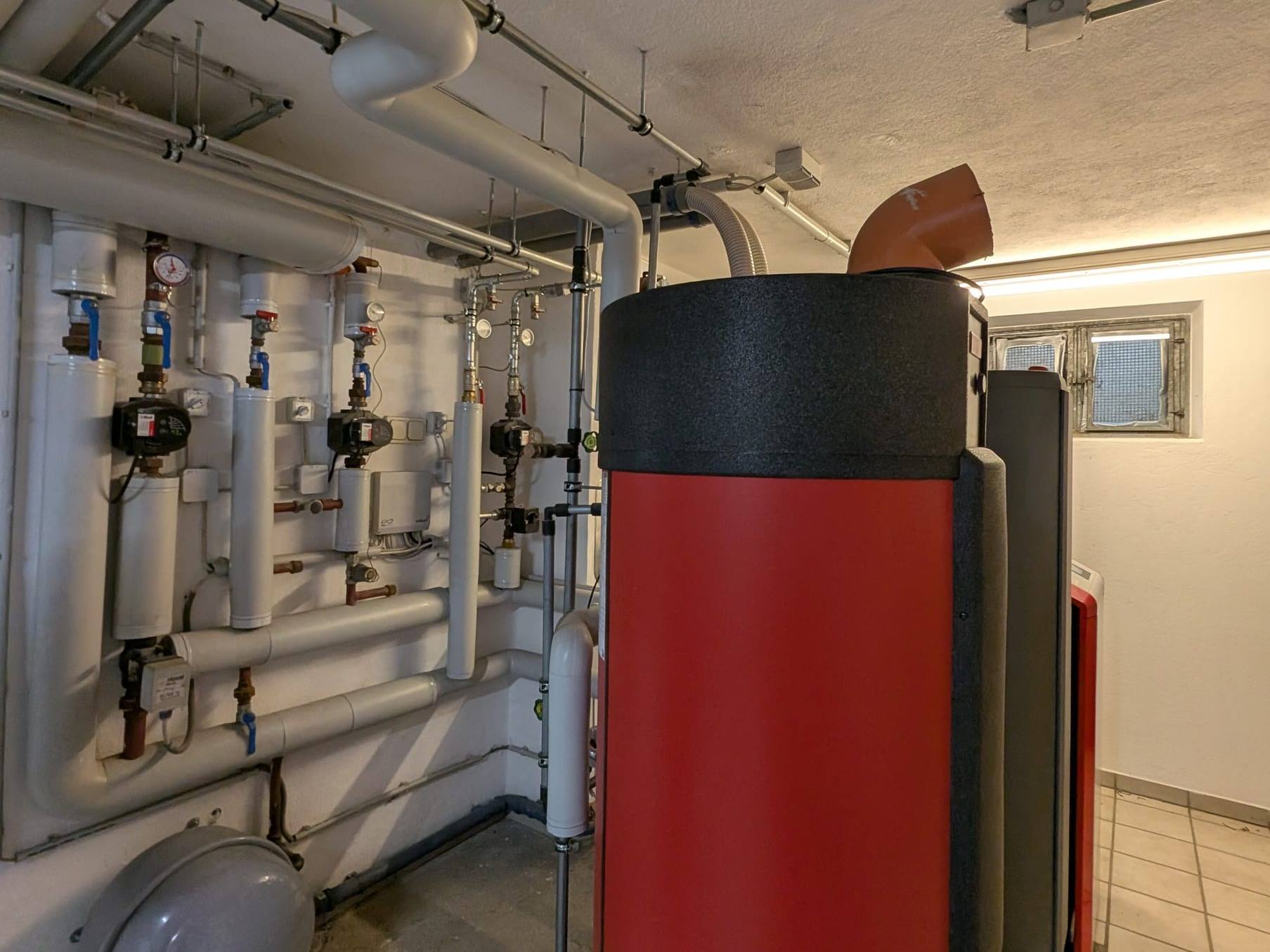 Heizraum mit gedämmten Rohrleitungen, Umwälzpumpen an der Wand und rotem Speicher