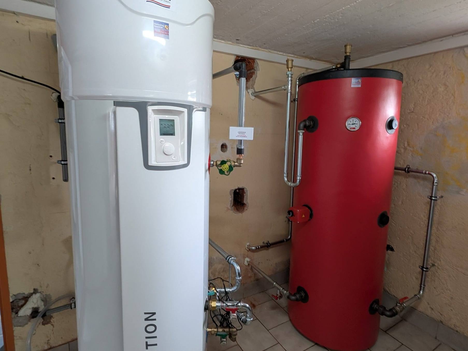 Weiße Warmwasser-Wärmepumpe mit der Aufschrift REVOLUTION neben einem roten Speicher im Keller