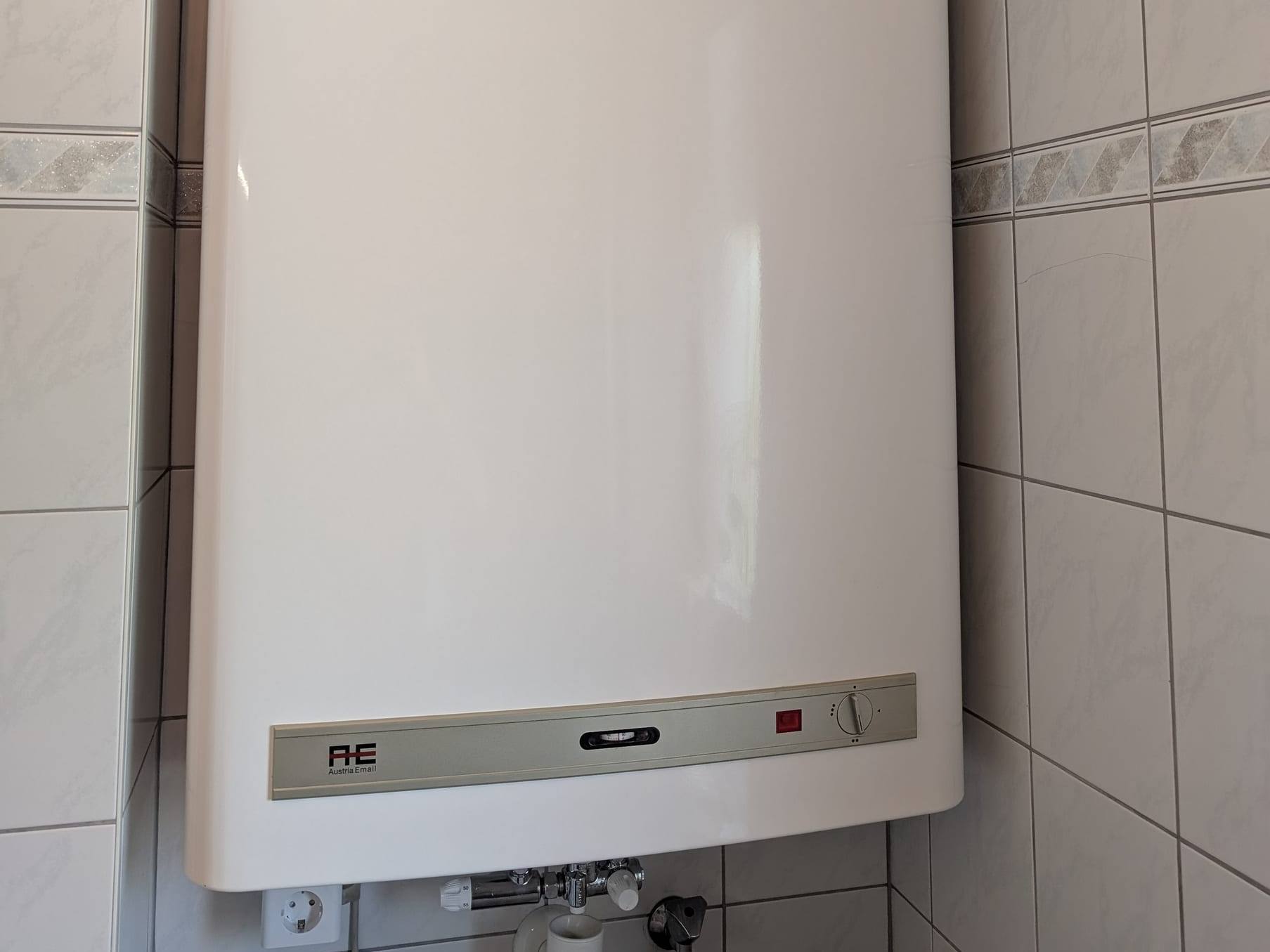 Weißer Wand-Warmwasserspeicher von Austria Email an gefliester Wand, darunter Anschlussarmaturen