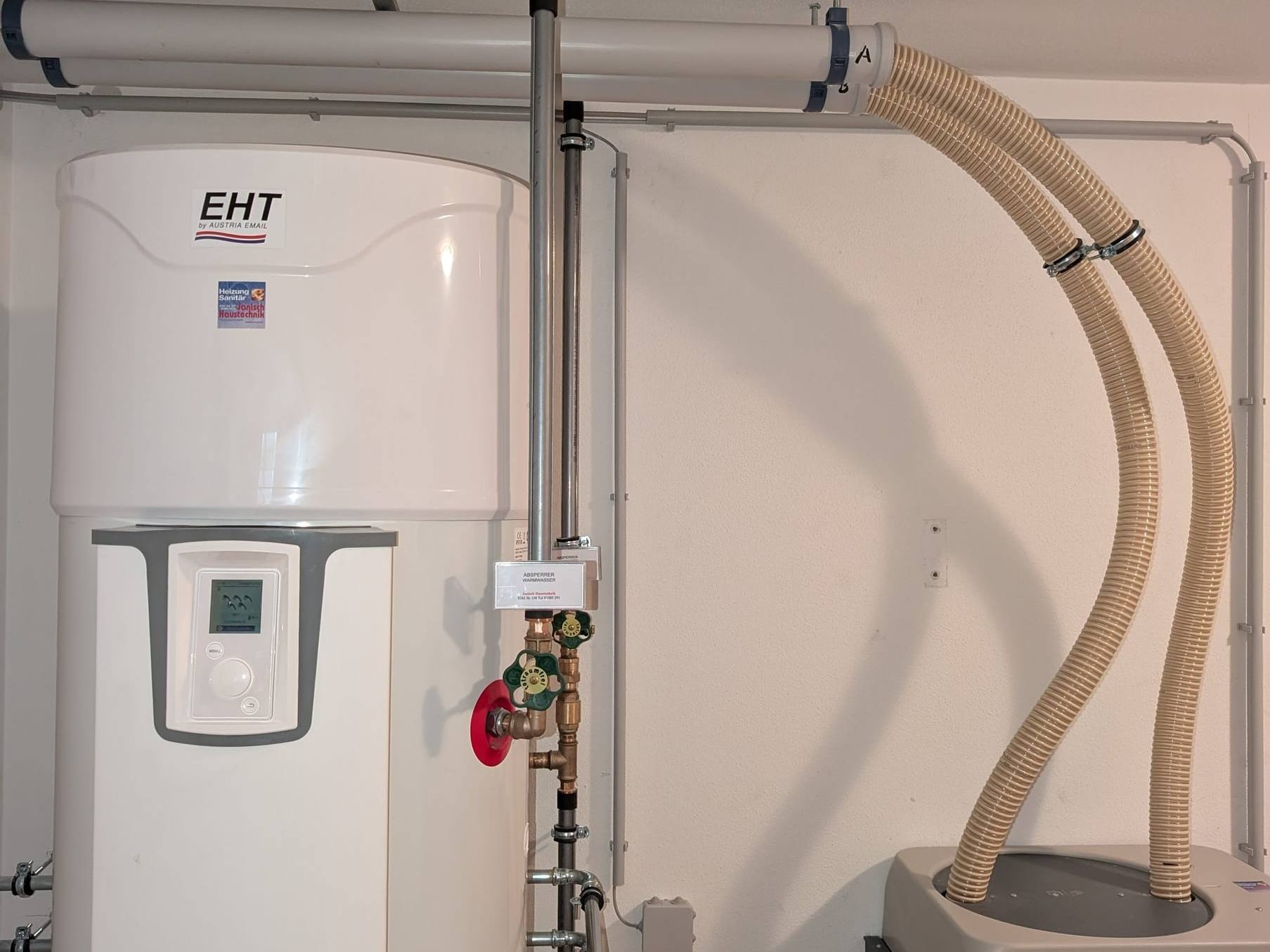 Weiße Warmwasser-Wärmepumpe mit Absperrventilen, daneben Pellets-Saugschläuche im Heizraum