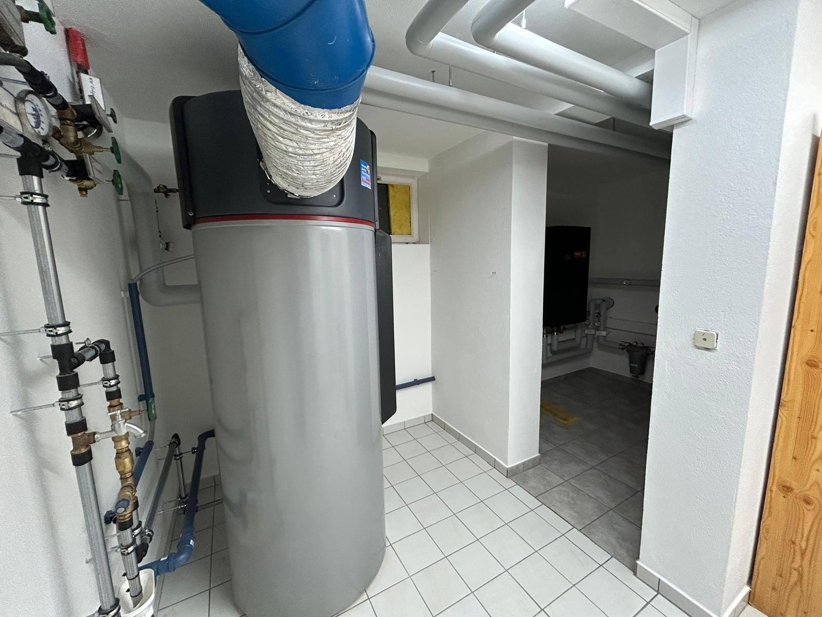Warmwasser-Wärmepumpe mit Anschluss an einen blauen Luftkanal in einem gefliesten Technikraum