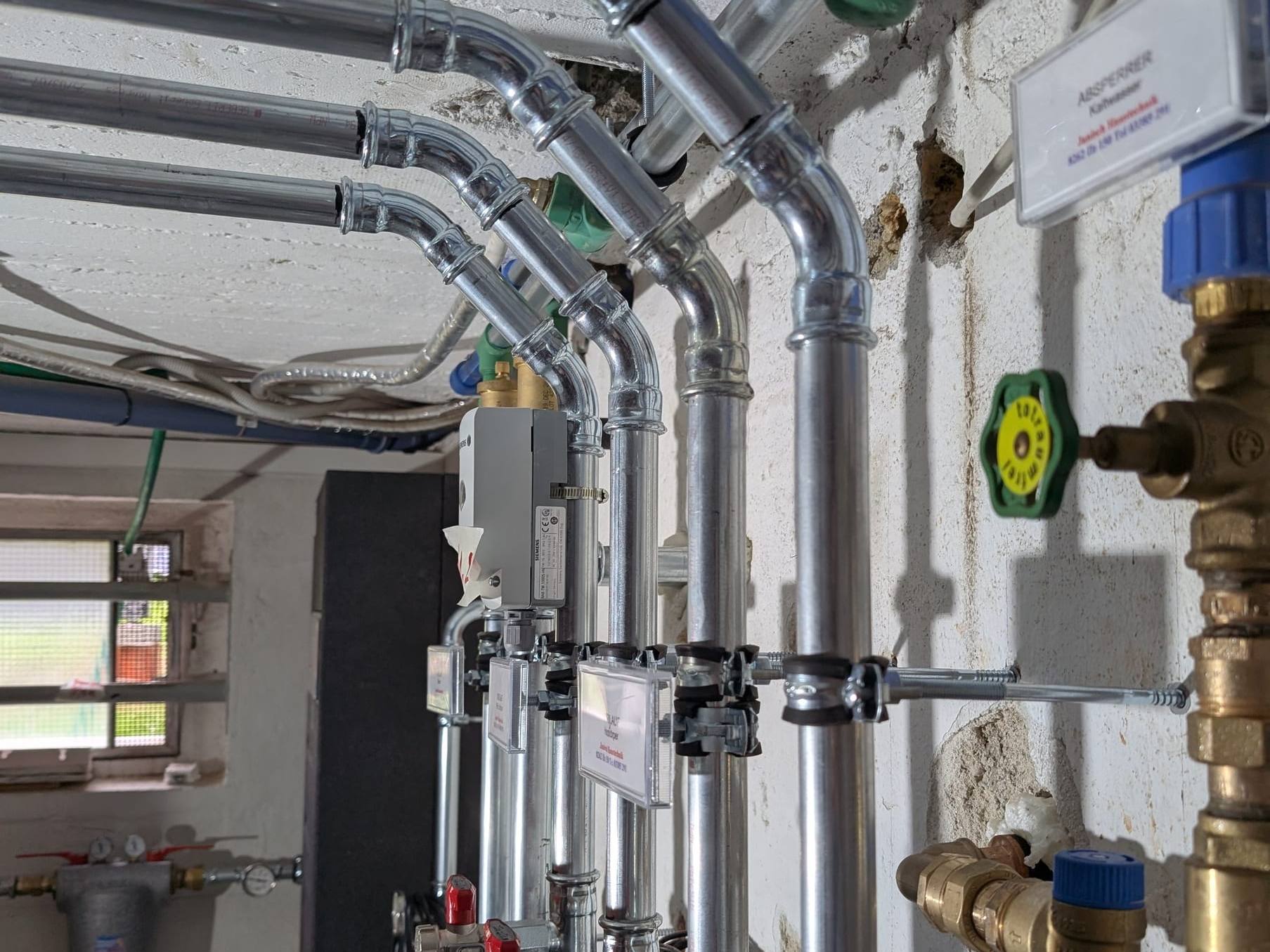 Parallel geführte Stahlrohre mit Pressfittings und beschrifteter Kaltwasser-Absperrer im Heizraum