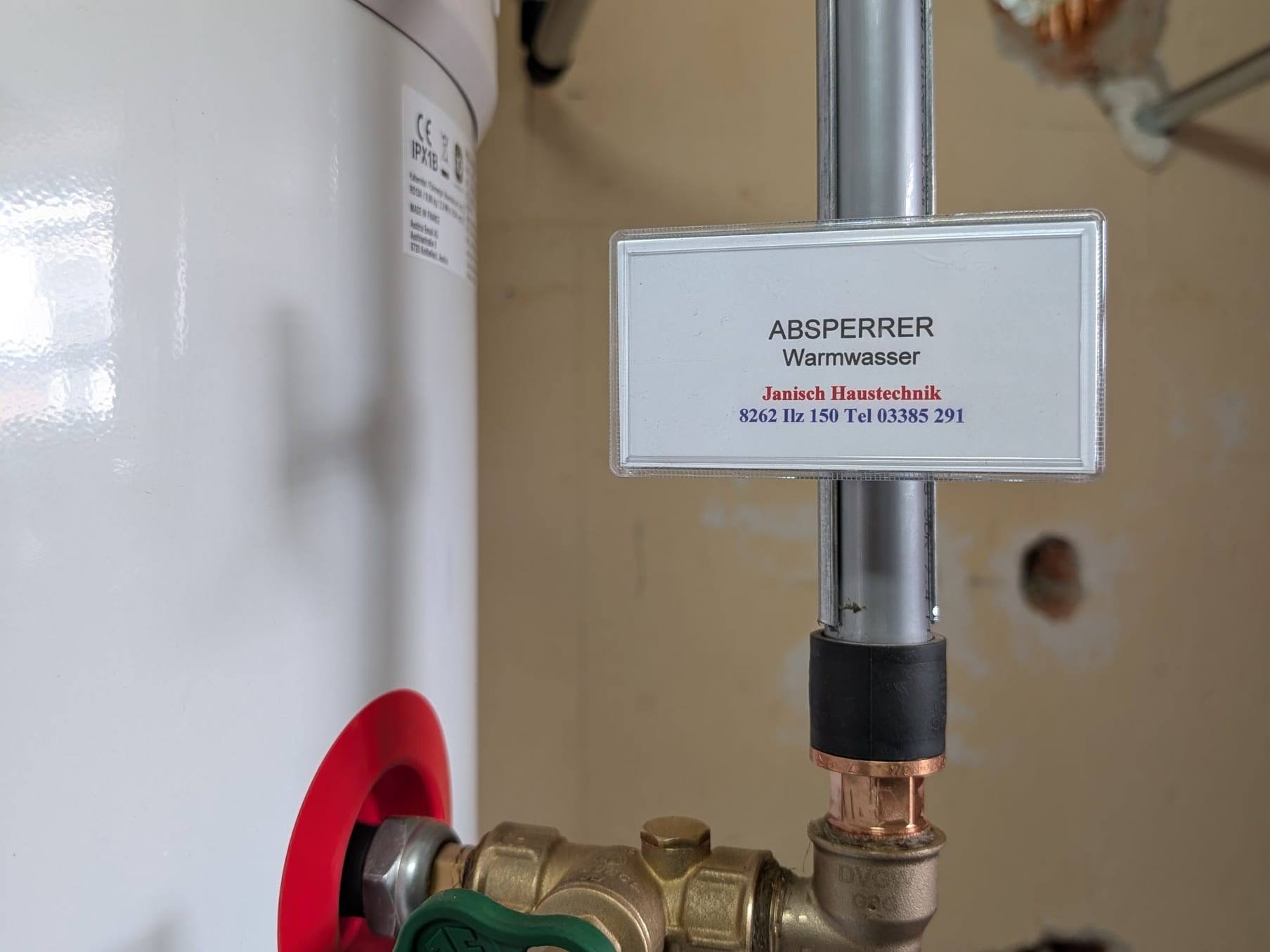 Grünes Absperrventil am Warmwasseranschluss mit dem Janisch-Schild „Absperrer Warmwasser“