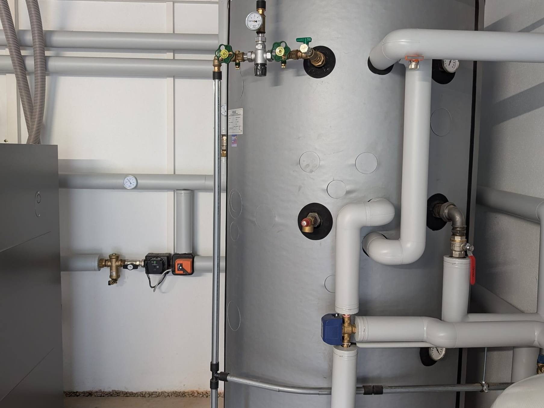 Grauer Speicher mit Thermometer, Armaturen und weiß gedämmten Anschlussleitungen im Heizraum