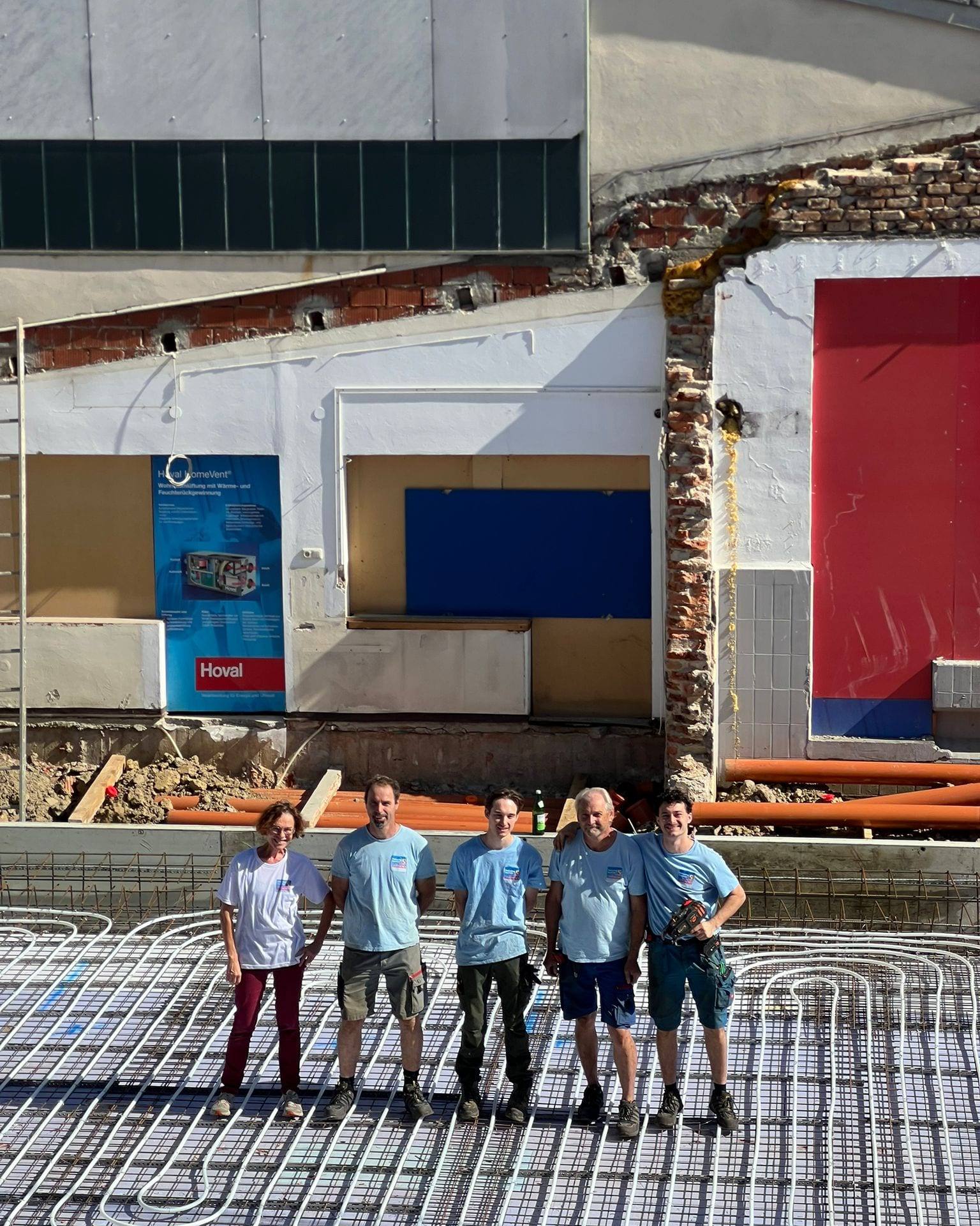 Fünf Personen in hellblauen Janisch-T-Shirts stehen auf einer Baustelle auf verlegten Heizungsrohren