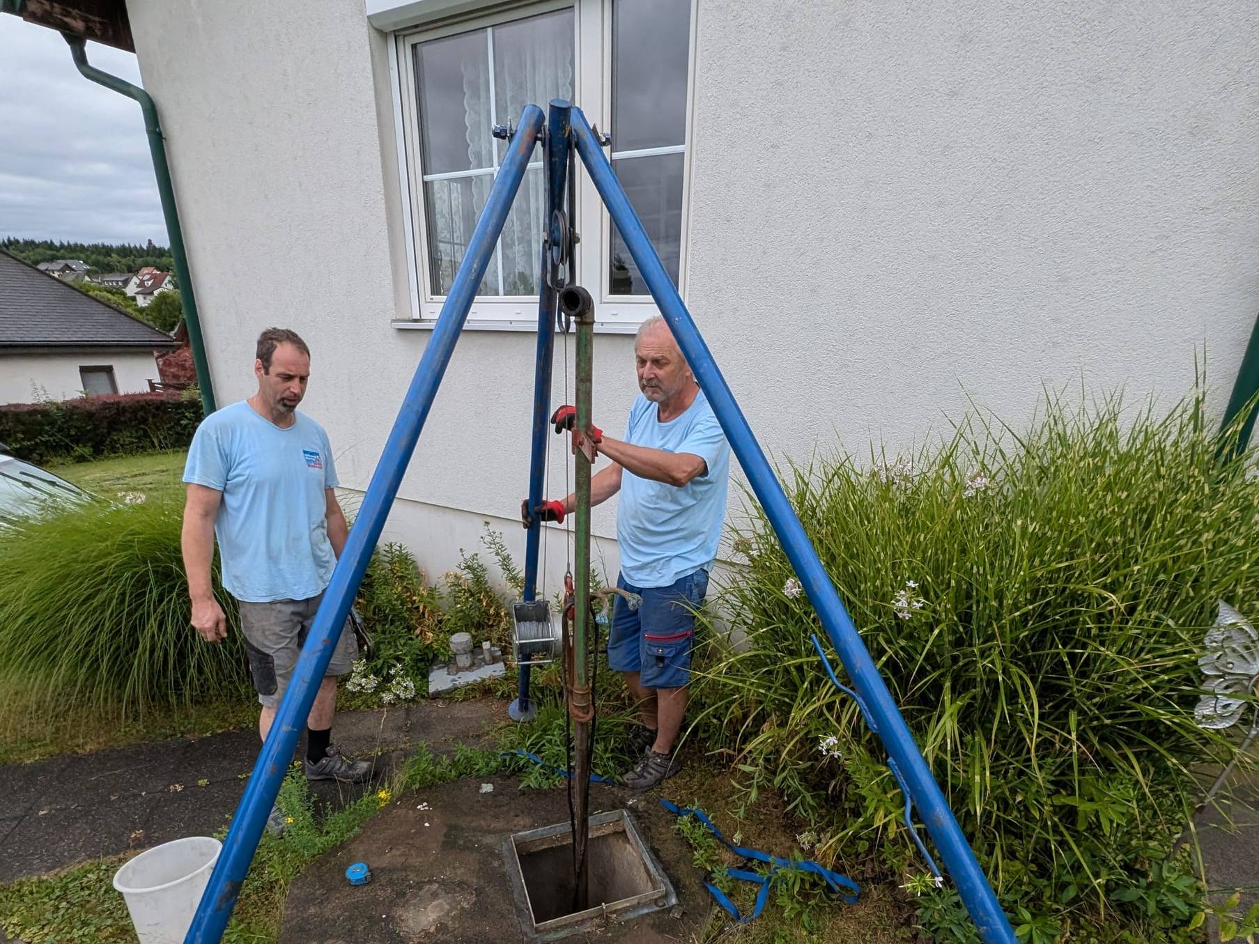 Zwei Monteure von Janisch Haustechnik setzen mit einem Dreibein und Seilzug eine Brunnenpumpe in einen Hausbrunnen