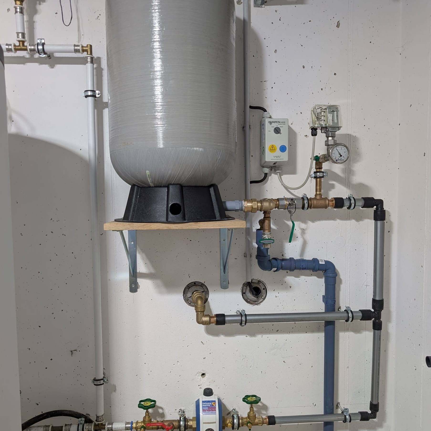 Grauer Druckkessel mit Pumpensteuerung, Druckschalter und Leitungen einer Brunnenwasseranlage an einer weißen Wand