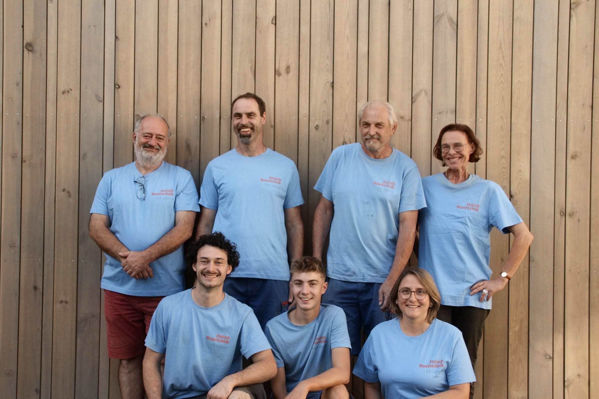 Das Team von Janisch Haustechnik in hellblauen T-Shirts „125 Jahre“ vor einer Holzfassade, alle lächeln in die Kamera