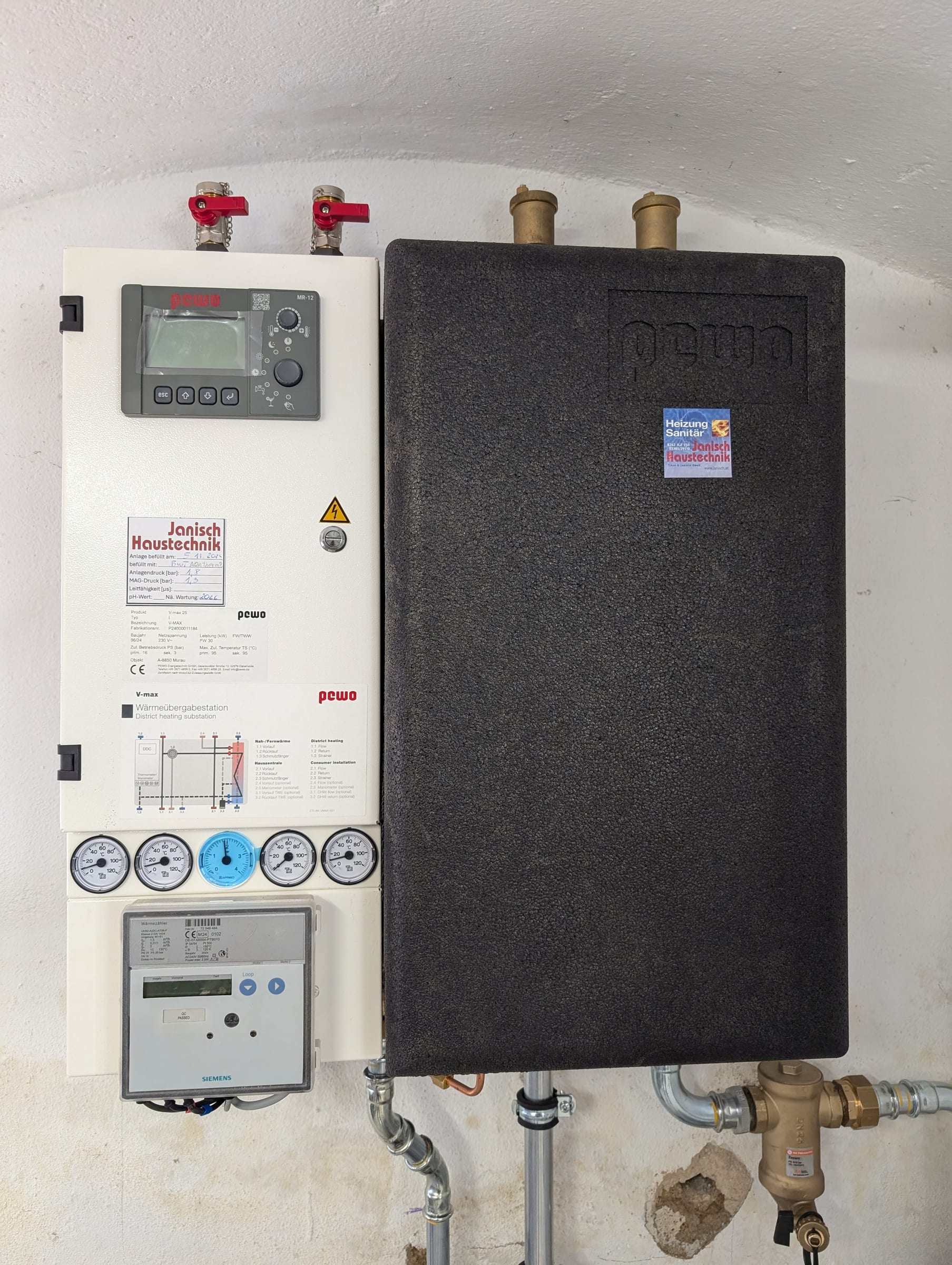 Wärmeübergabestation mit Regler, Thermometern, Wärmemengenzähler und Janisch-Aufkleber an einer Kellerwand