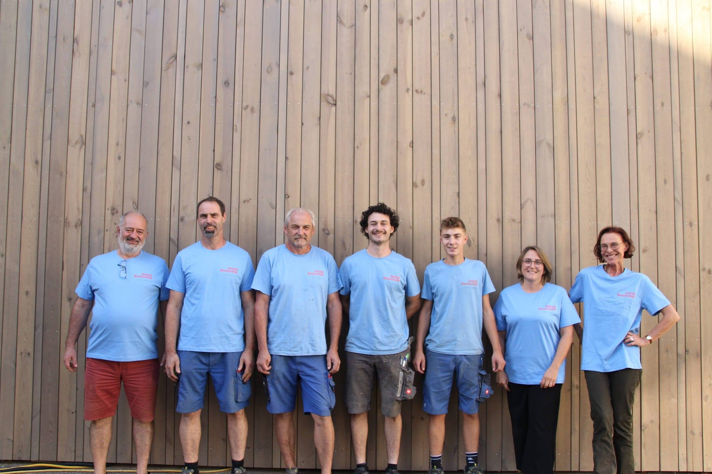 Das Team von Janisch Haustechnik in hellblauen Firmen-T-Shirts nebeneinander vor einer Holzfassade